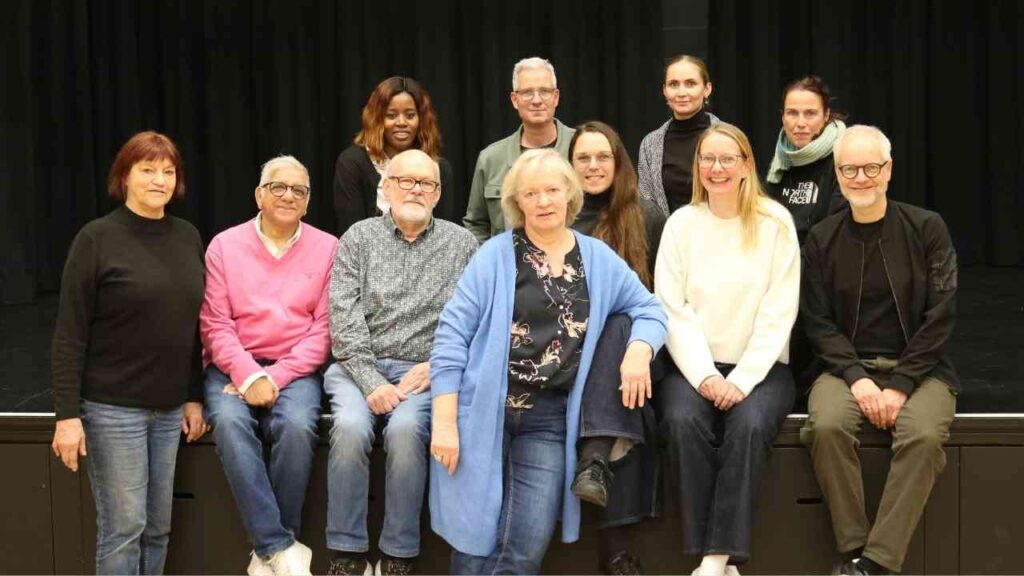 Gruppe von Personen, die auf einer Bühne in einem Theater oder Veranstaltungsort posieren, mit unterschiedlichen Altersgruppen und Geschlechtern.