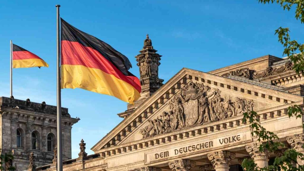 Das Reichstagsgebäude in Berlin mit der deutschen Flagge im Vordergrund und dem Schriftzug 'Dem Deutschen Volke'.