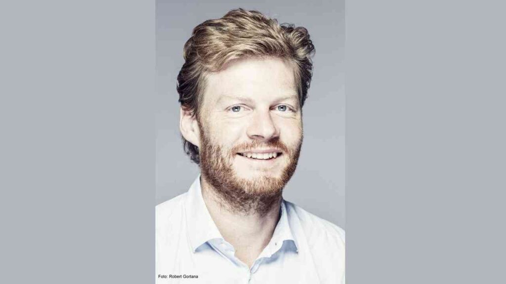 Porträt eines Mannes mit blonden Haaren und Bart, der in die Kamera lächelt, in einem hellen Hemd.