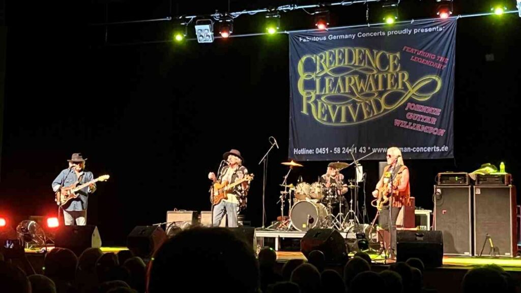 Creedence Clearwater Revived Bandmitglieder spielen auf der Bühne während eines Konzerts mit einem Banner im Hintergrund.