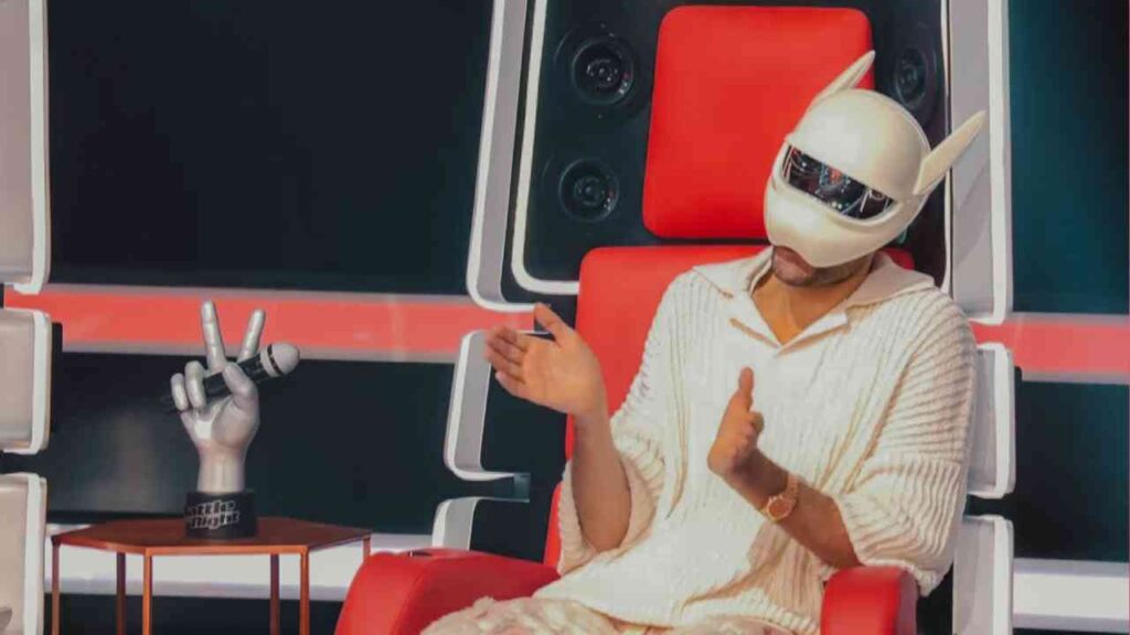 Person mit einer weißen Maske und einem roten Pullover sitzt auf einem roten Stuhl bei einer Talentshow.