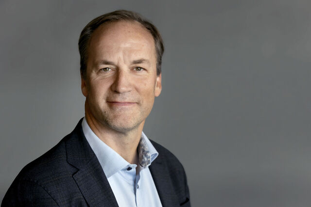 Porträt eines Mannes in einem dunklen Anzug mit einem hellen Hemd, lächelnd vor einem grauen Hintergrund.