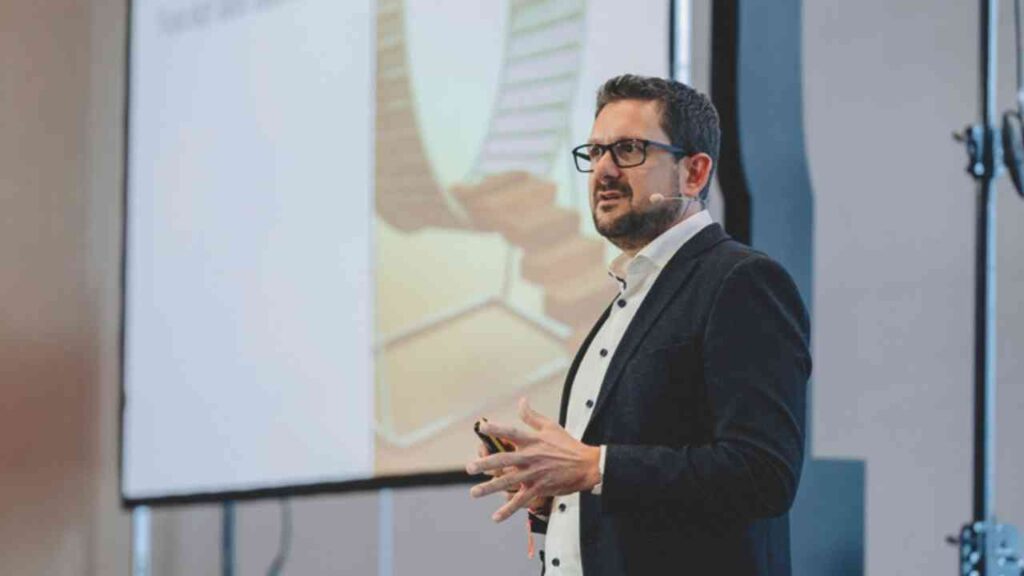 Mann mit Brille und Mikrofon spricht auf einer Bühne während einer Präsentation zu einem Publikum.