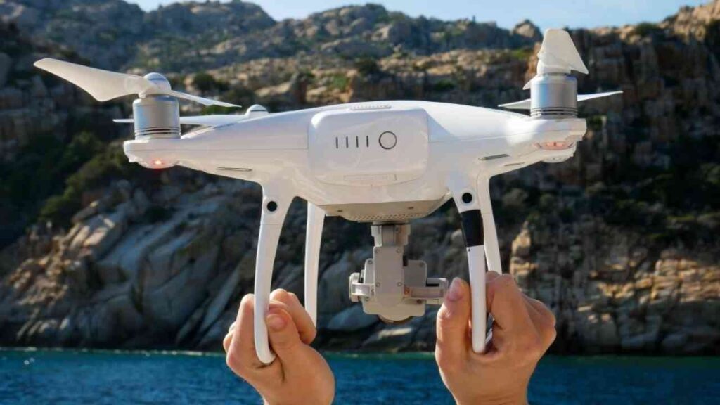 Weißer Quadrokopter wird von einer Person über Wasser gehalten, mit Felsen im Hintergrund.