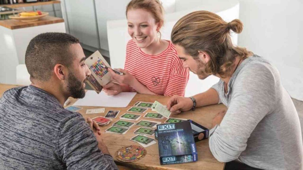 Drei Personen spielen ein Brettspiel am Tisch mit Karten und Spielmaterialien.