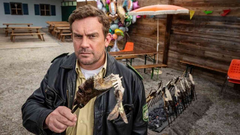 Mann in Lederjacke hält einen Spieß mit gegrilltem Fisch in einer Außenumgebung mit Holzstruktur und bunten Dekorationen.