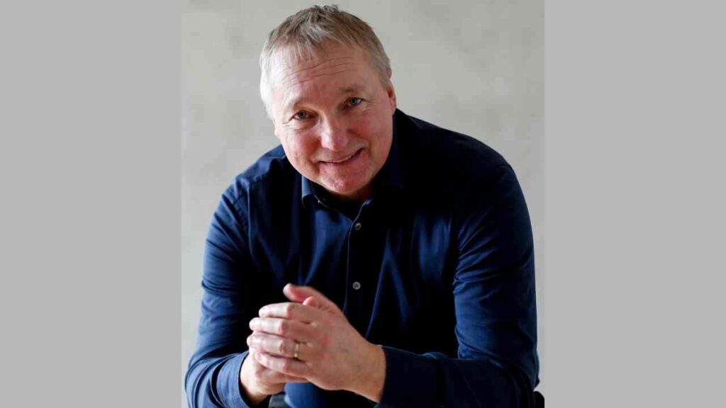 Mann in dunkelblauem Hemd lächelt freundlich in die Kamera, vor grauem Hintergrund sitzend.