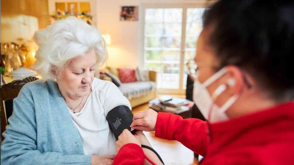 Ältere Frau erhält Blutdruckmessung von einer Pflegekraft in einem Wohnraum.