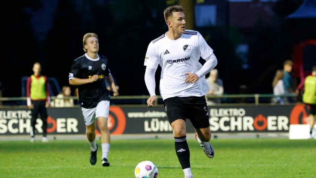 Fußballspieler in einem weißen Trikot mit schwarzem Logo, der den Ball während eines Spiels führt.