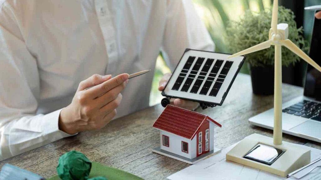 Person hält ein Solarpanel und zeigt auf ein Modellhaus und eine Windkraftanlage auf einem Tisch.