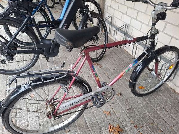 Rotes Fahrrad mit einem schwarzen Sattel und einem Gepäckträger, abgestellt in einer Fahrradabstellanlage.