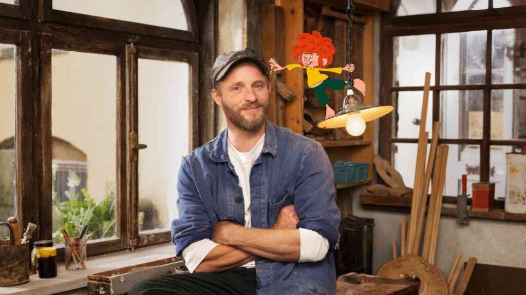 Florian Brückner sitzt in einem Raum mit Holzarbeiten, während Pumuckl über einer Lampe schwebt.