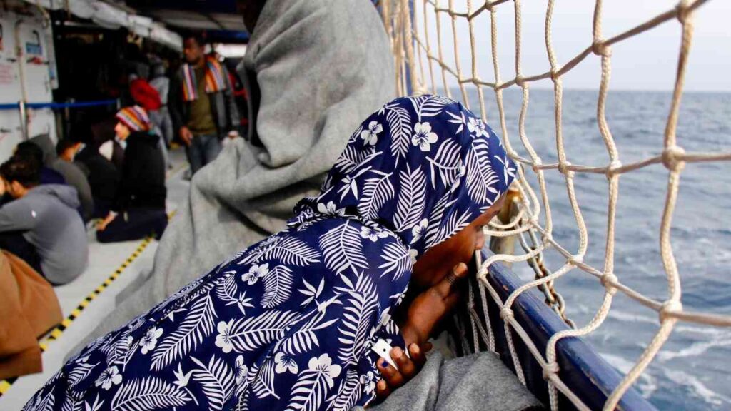 Person in blauer Kleidung mit floralem Muster, die über die Reling eines Bootes auf das Meer schaut.
