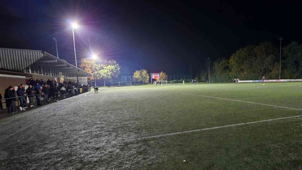 Flutlichtanlage auf einem Kunstrasenplatz mit Zuschauern am Rand des Spielfelds bei Nacht.