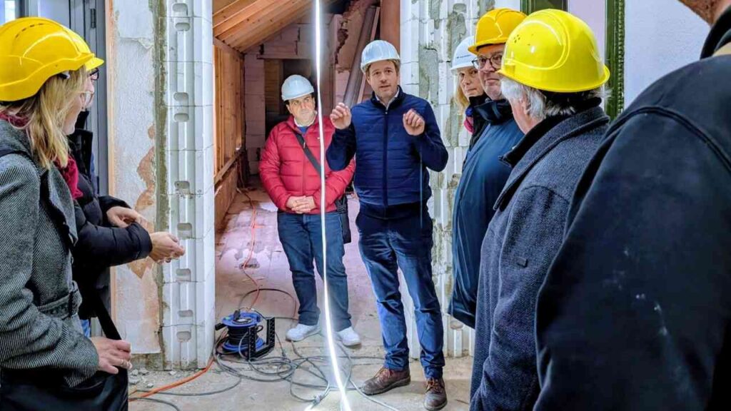 Gruppe von Personen mit Sicherheitshelmen, die sich in einem Bauprojekt der Musikschule versammeln und einem Sprecher zuhören.