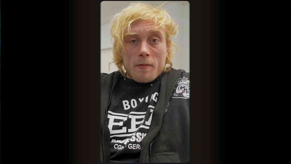 Mann mit blonden Haaren und schwarzem T-Shirt, das den Text 'Boxing' zeigt, in einem Innenraum.