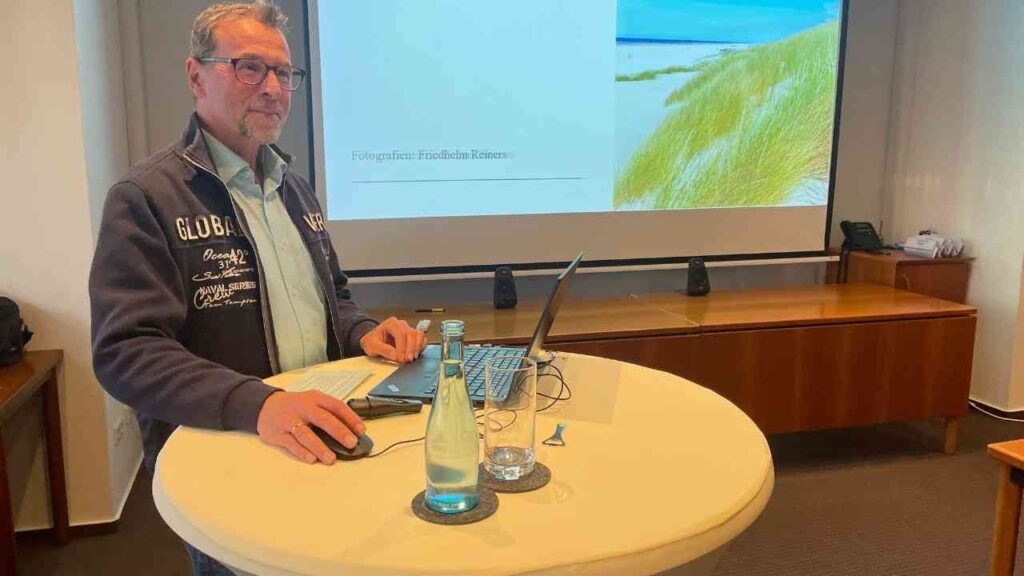 Mann steht an einem Stehpult mit Laptop, Wasserflasche und Präsentation im Hintergrund.