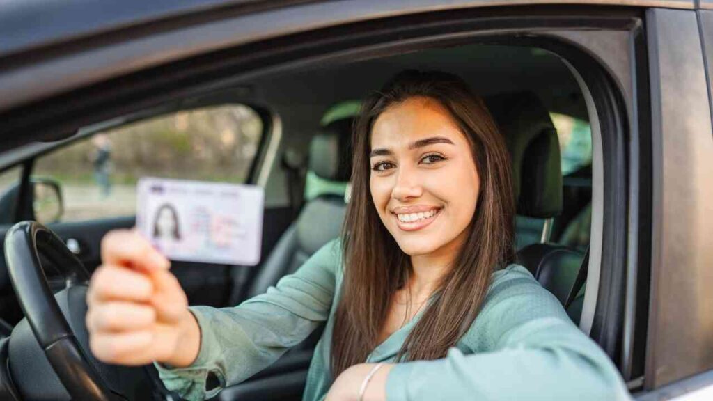 Junge Frau hält ihren Führerschein aus dem Autofenster und lächelt in die Kamera.