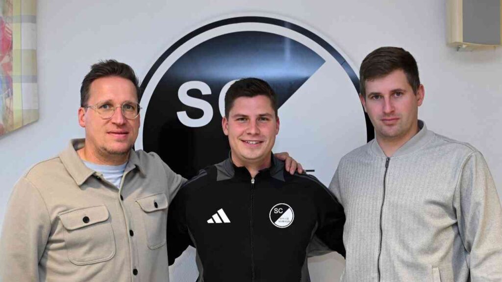 Drei Männer stehen zusammen vor einem SC Spelle-Venhaus Logo, einer trägt einen schwarzen Trainingsanzug.