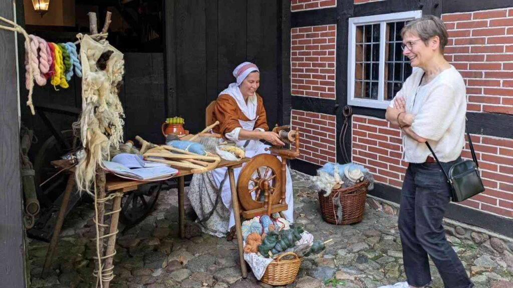 Frau mit Spinnrad, die Wolle spinnt, während eine Zuschauerin zuschaut, in einem historischen Handwerksumfeld.