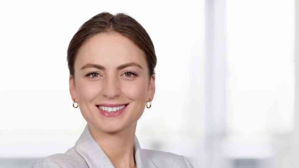 Frau mit braunen Haaren und Ohrringen lächelt in einem hellen, professionellen Umfeld.