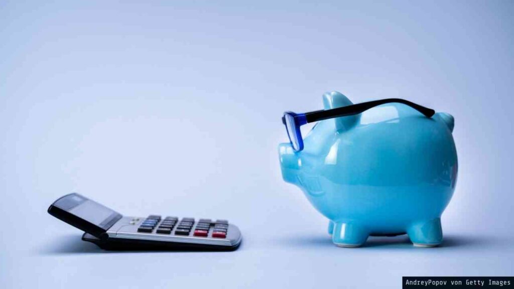 Blauer Sparschwein mit Brille neben einem Taschenrechner auf einem hellen Hintergrund.