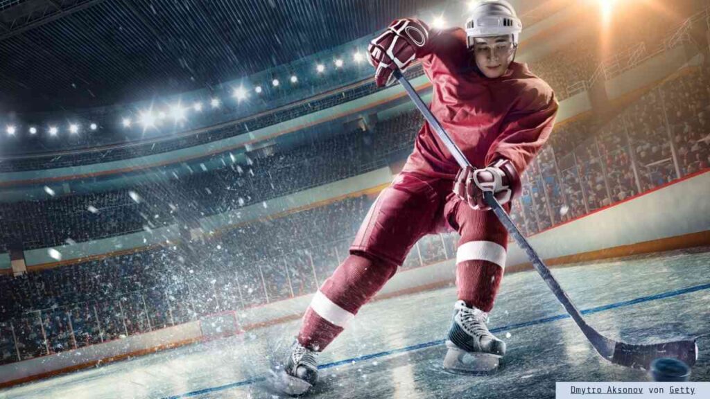 Eishockeyspieler in rotem Trikot, der auf dem Eis in einer Arena spielt und den Puck mit einem Schläger führt.