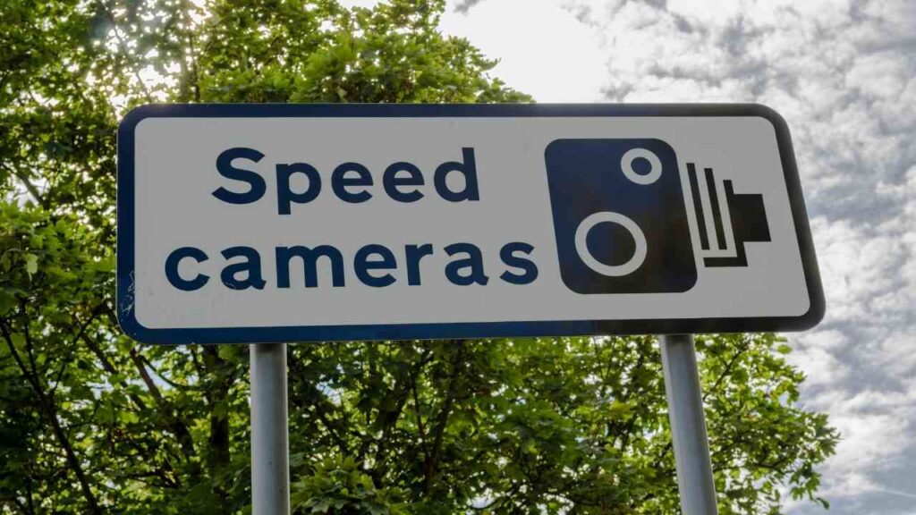 Verkehrsschild mit der Aufschrift 'Blitzgeräte' und einem Symbol einer Kamera, aufgestellt in einer grünen Umgebung.