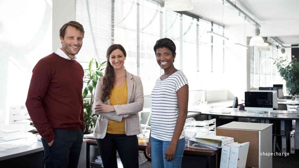 Drei Personen in einem modernen Büro, die freundlich in die Kamera lächeln und sich in einem kreativen Arbeitsumfeld befinden.