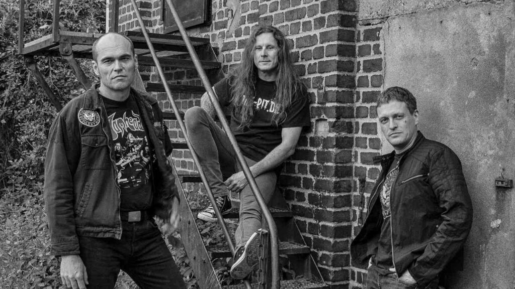 Drei Männer posieren in einer urbanen Umgebung, zwei stehen und einer sitzt auf einer Treppe.