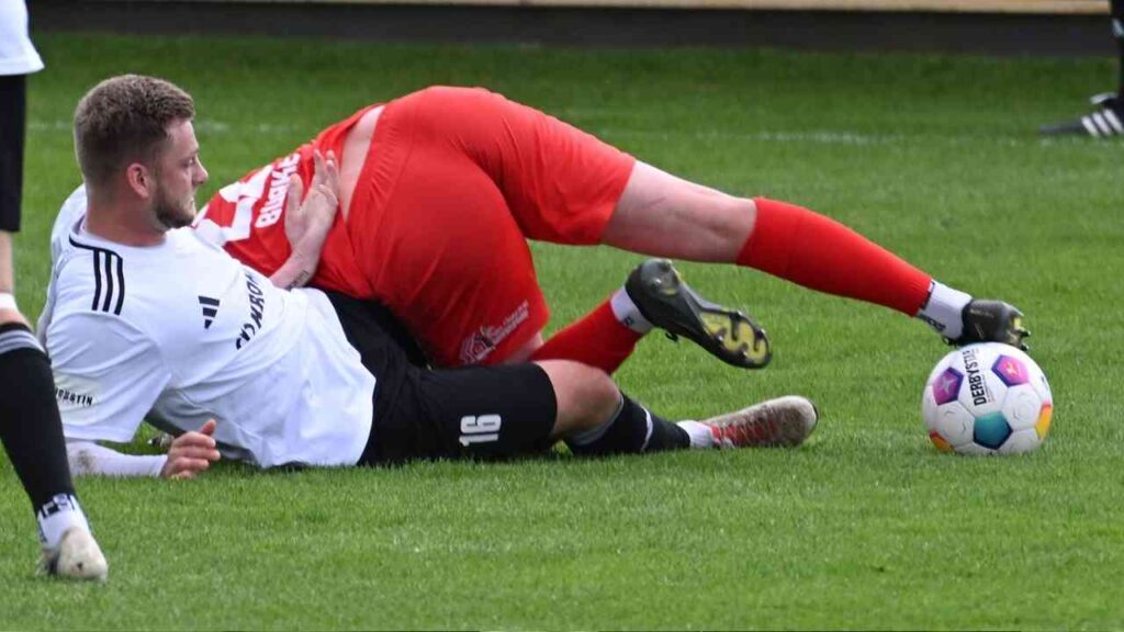 Zwei Fußballspieler im Wettkampf auf dem Spielfeld, einer liegt auf dem Boden und der andere ist darüber gebeugt.