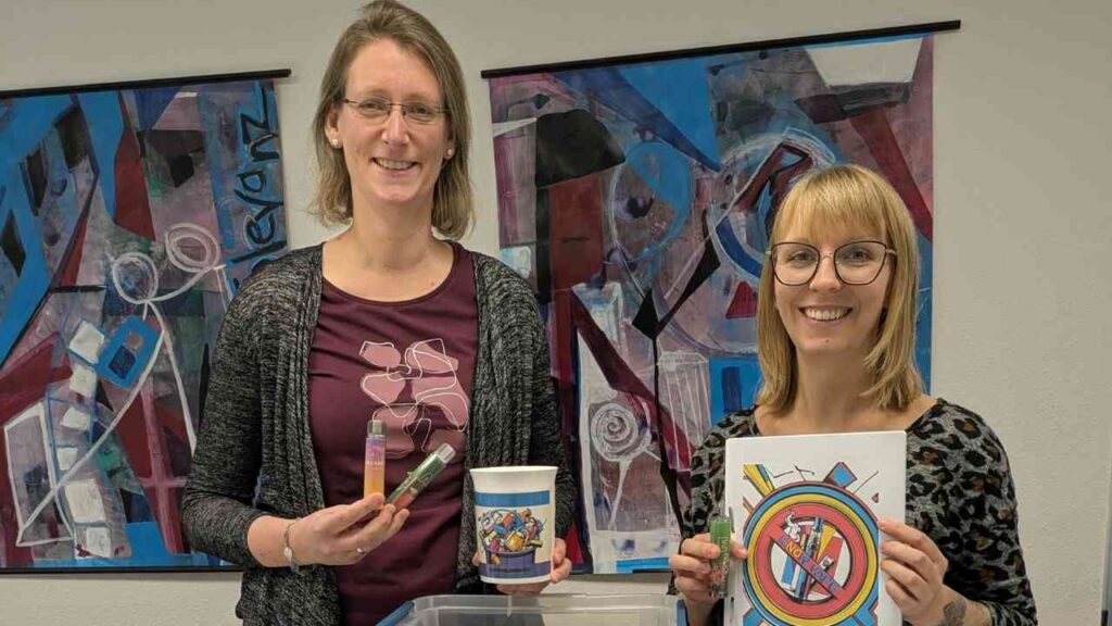 Zwei Frauen halten verschiedene Gegenstände, darunter Marker und eine bunte Zeichnung, vor einem abstrakten Kunstwerk.