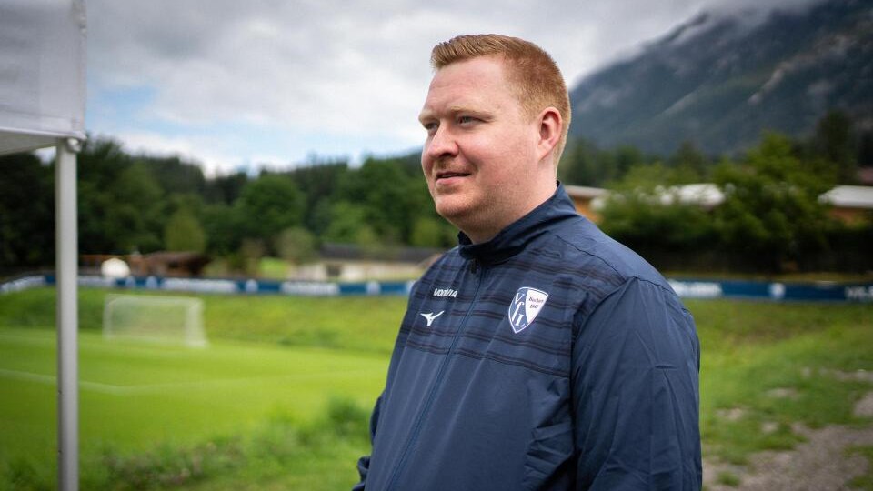 Mann in Sportjacke steht auf einem Fußballfeld mit grüner Wiese im Hintergrund.