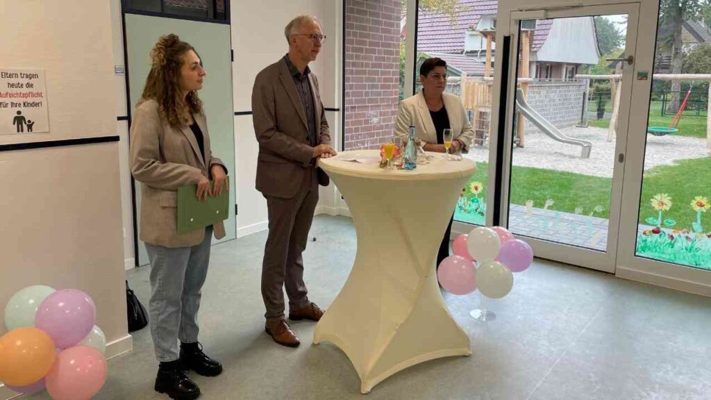 Drei Personen stehen an einem Stehtisch in einem Kindergarten mit bunten Luftballons im Hintergrund.