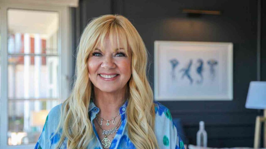 Frau mit langen blonden Haaren, lächelnd, trägt ein blaues Hemd mit bunten Mustern und Schmuck.