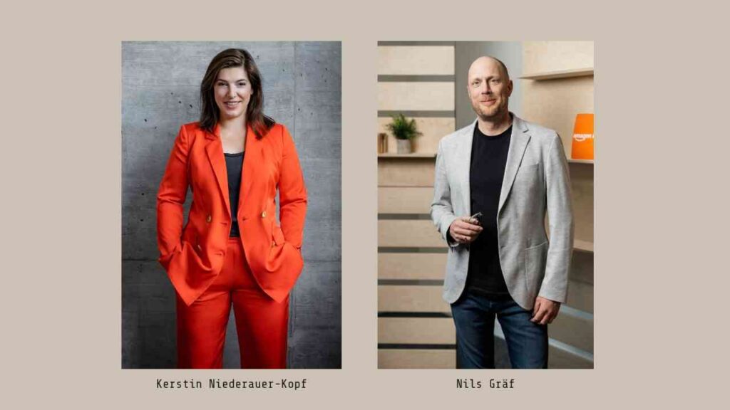 Kerstin Niederauer-Kopf in einem orangefarbenen Anzug und Nils Graef in einem grauen Blazer, beide stehen vor neutralem Hintergrund.