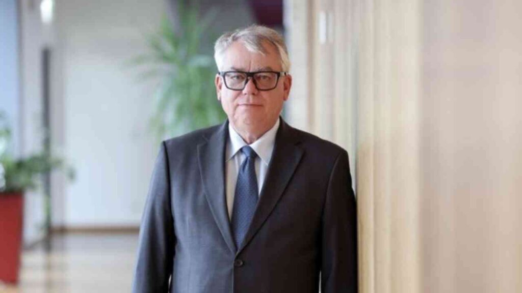 Ein älterer Herr im Anzug steht in einem Bürogebäude neben einer Holzwand.