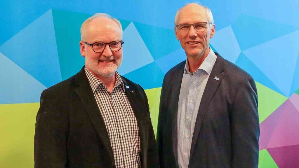 Zwei Männer in Anzügen vor einem bunten geometrischen Hintergrund lächeln in die Kamera.