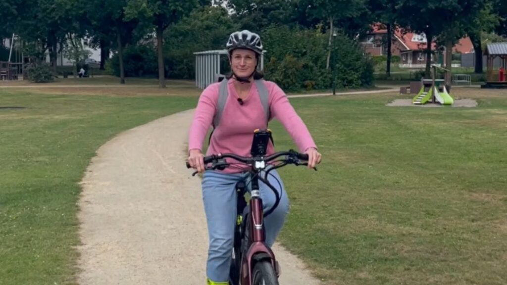 Frau auf einem Fahrrad in einem Park, trägt einen Helm und hat ein Mikrofon am Kragen.