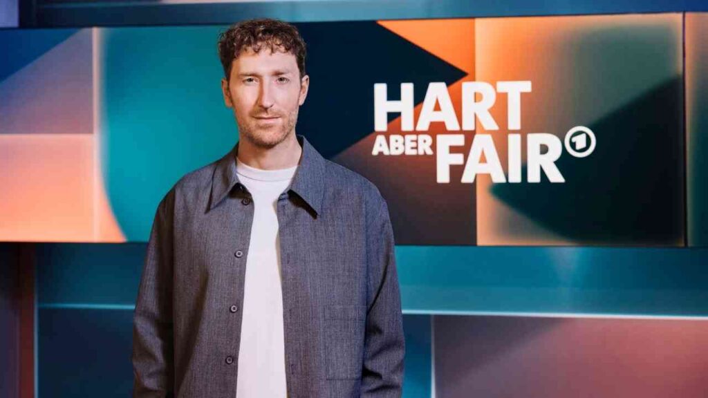 Mann mit lockigem Haar in einem grauen Jackett vor einem bunten Hintergrund mit dem Schriftzug 'HART ABER FAIR'.