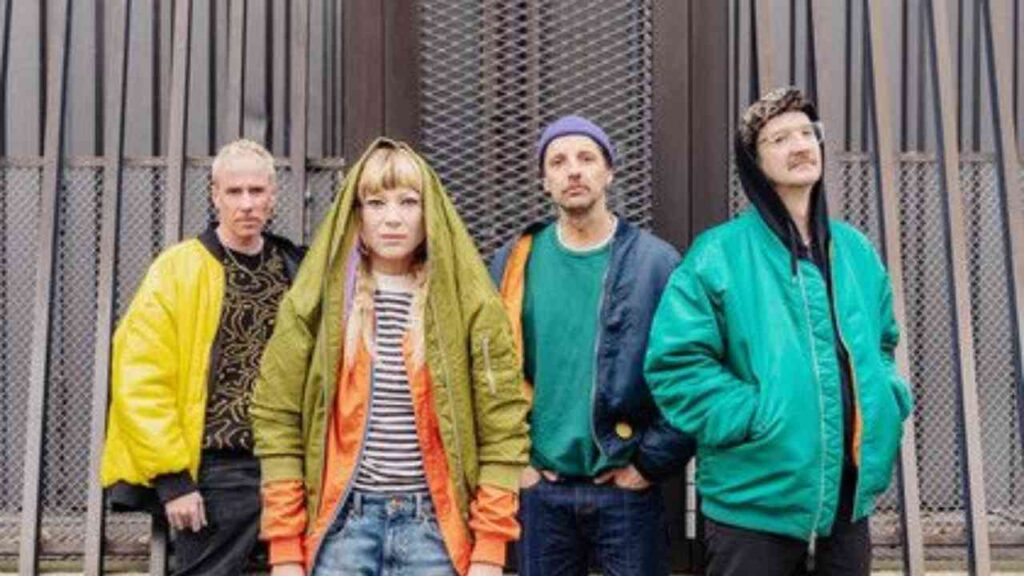 Vier Personen in bunten Jacken stehen vor einem Gittertor in urbaner Umgebung.