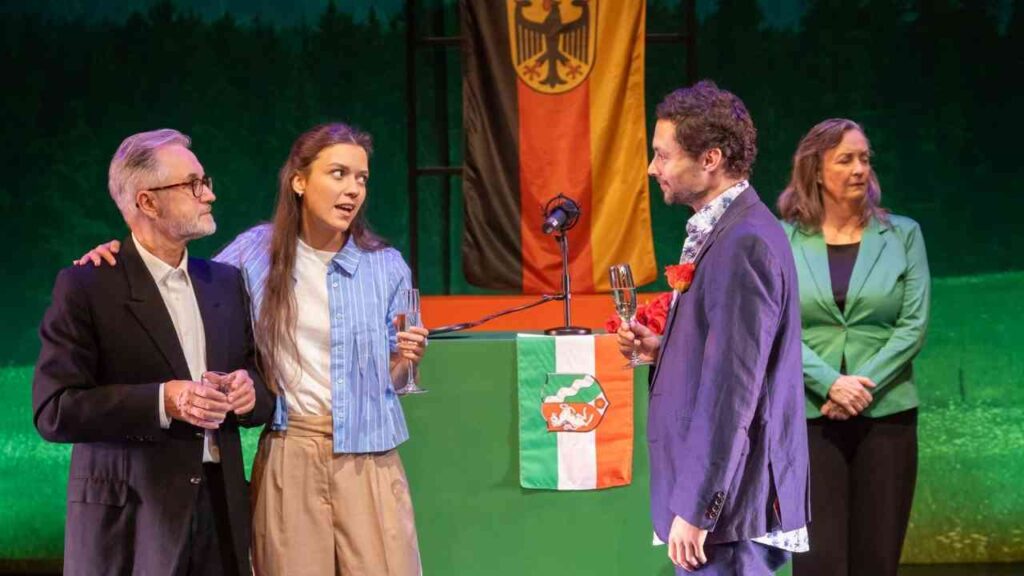 Schauspieler auf der Bühne während einer Theateraufführung mit einem deutschen Flaggenhintergrund.