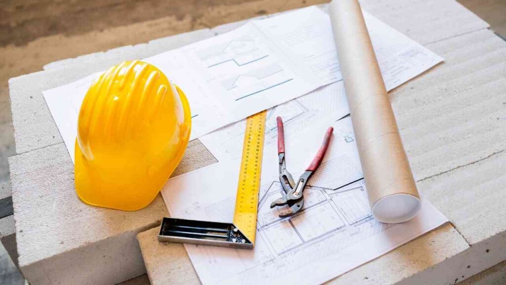 Gelbe Bauhelm, Baupläne, Zollstock und Zange auf einem Tisch aus Baustellenmaterial.