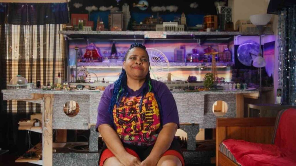 Person mit bunten Haaren sitzt vor einer detailreichen Modelllandschaft mit Stadtansicht und Attraktionen.