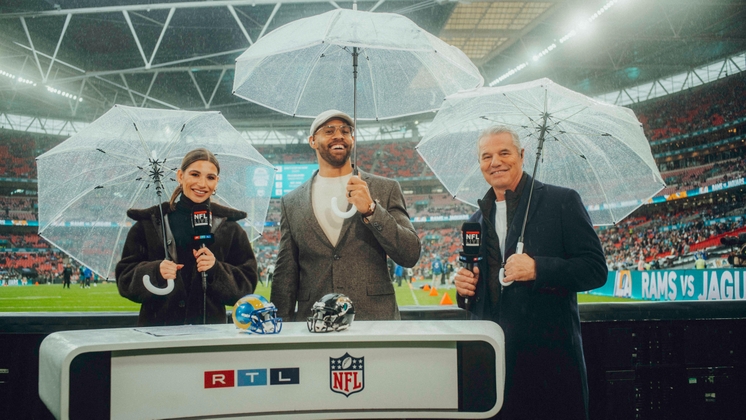 Drei Moderatoren mit Regenschirmen stehen am Kommentatorenpult während eines NFL-Spiels im Wembley-Stadion.