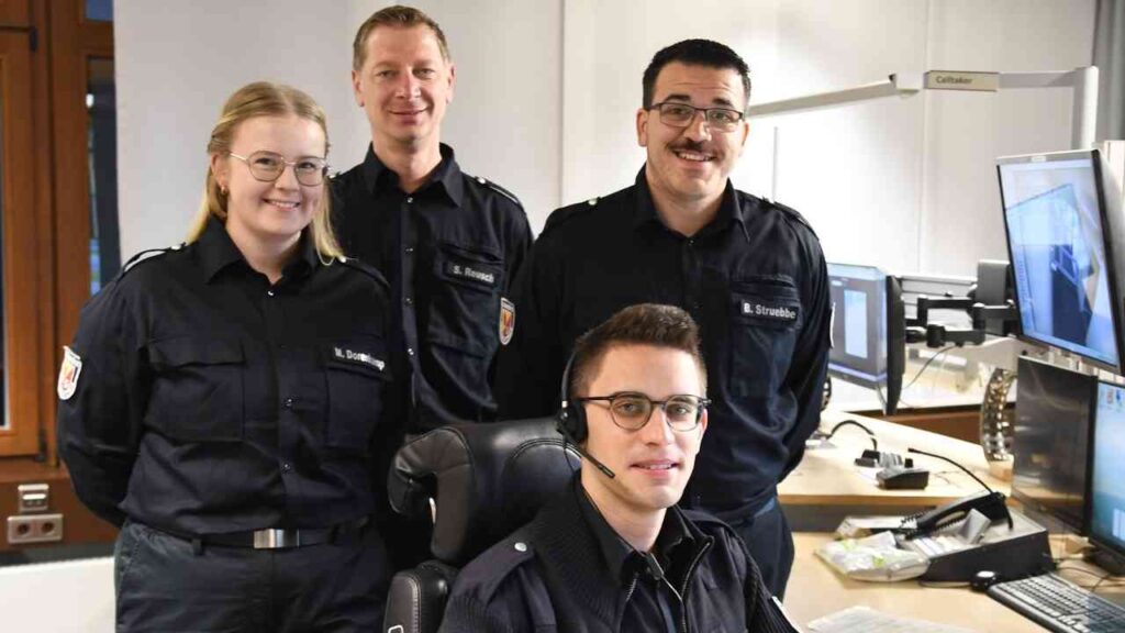 Vier Personen in Uniform, die in einem Büro für Notrufdienste arbeiten, mit einem Mitarbeiter am Computer und Headset.