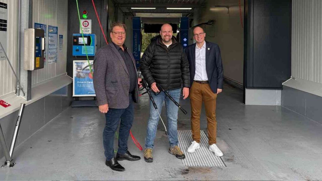 Drei Männer stehen in einer Autowaschanlage, einer hält einen Hochdruckreiniger in der Hand.