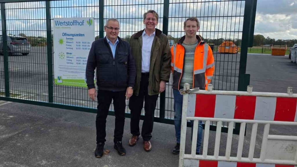 Drei Personen stehen vor dem Eingang eines Wertstoffhofs mit einem Informationsschild über Öffnungszeiten.