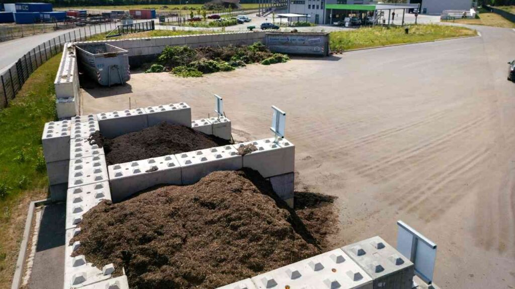 Haufen von organischem Abfall auf einem Recyclinggelände mit Betonmauern und einem Container im Hintergrund.