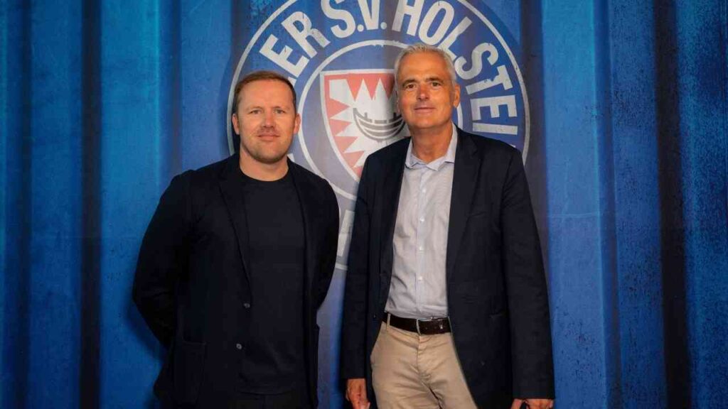 Zwei Männer stehen vor Holstein Kiel Logo, blauem Hintergrund, Business-Kleidung.